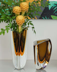 A Gardeco Glass Vase Triangle 3 Fume Amber with contemporary refinement, holding a bouquet of flowers and a separate Gardeco triangular vase filled with leaves.