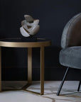 Abstract sculpture on a round table next to a gray armchair against a dark wall.