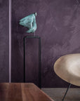 Bronze sculpture on a stand against a textured purple wall with a wooden table and chair in the foreground.