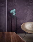 Bronze sculpture on a stand against a textured purple wall with a wooden table and chair in the foreground.
