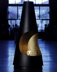 A modern black outdoor fireplace with a sculptural cone shape and a curved cutout, shown lit with a warm fire inside. The background is a dark, atmospheric indoor space with blurred figures and large windows.