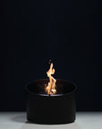 A modern black cylindrical bioethanol fireplace with a visible orange and yellow flame, set against a dark studio background.