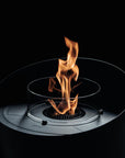 A modern circular outdoor fire pit with a clear glass wind guard and a central flame, set against a solid black background.