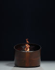 A modern cylindrical indoor bioethanol fire pit with a dark copper wood-grain finish, featuring a clear glass wind guard and a small flame burning inside, set against a dark studio background.