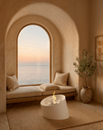 A serene coastal nook with a large arched window overlooking the ocean at sunset, featuring a cushioned bench, pillows, a small white sculptural table with a flame, a potted olive tree, and warm plaster walls.