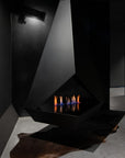 A modern black geometric wall-mounted fireplace with a reflective glass firebox showing orange and blue flames, set in a dark minimalist room with concrete floors and a brown hide rug.