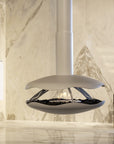 A modern floating fireplace with a white ceramic or stone-look outer shell, suspended over black decorative stones with a visible flame, set against a light beige marble wall and floor.