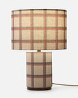 A table lamp with a cylindrical base and matching drum shade, both upholstered in a beige plaid fabric with red and dark blue stripes, shown on a light gray background.