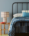 A styled bedroom scene featuring a black metal bed frame with a blue and cream patterned quilt, striped pillows, a wooden side table, and a plaid table lamp against a muted blue-gray wall.