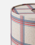 Close-up of a cylindrical lampshade or fabric-covered container corner, showing a woven plaid pattern in beige, red, and blue on a light gray background.