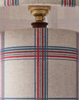Close-up of a cylindrical table lamp shade with a woven beige fabric and a red and blue plaid pattern, showing part of the lamp's brass socket and wooden collar.