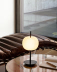Modern table lamp on a glass table with a brown cushioned chair in the background.