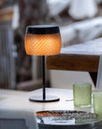 Table lamp with woven design on a table with glasses and a plate in a blurred outdoor setting