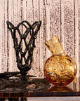 Decorative black metal sculpture and gold glass vase on a reflective surface with a textured wall background.