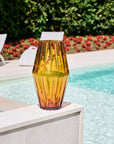 Amber crystal vase on a ledge by a pool with greenery and flowers in the background