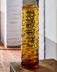Tall amber crystal vase on a wooden surface with a white brick wall background