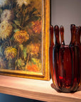 Brown crystal vase on a wooden shelf with a floral painting in the background