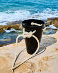 Black crystal vase tied with rope on a rocky surface with ocean waves in the background