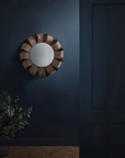 Round wooden mirror on a dark blue wall with a plant to the left.