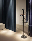 Black coat stand with a round base and a rectangular top in a room with dark curtains and tiled wall.