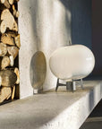 A modern white spherical table lamp with a small dark metal base, displayed on a stone mantelpiece next to a stack of firewood logs.