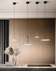Four modern linear pendant lights with black cords and gold-toned metal accents, hanging in a row above a kitchen island against a wood-paneled wall.