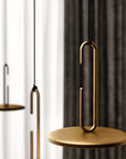 A modern pendant light with a matte gold circular disc shade and matching U-shaped metal frame, suspended by a thin black cord against a softly blurred background of white and dark gray curtains.