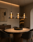 Three modern pendant lights with clear glass shades and warm glowing bulbs hang over a round wooden dining table in a stylish, warmly lit dining room with gray upholstered chairs.