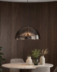 A modern semi-dome pendant light with a dark smoked glass shade and a warm glowing geometric metal interior, hanging above a dining table in a contemporary room with dark wood paneling.