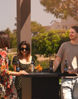 Four people standing around a gas fireplace table with drinks outdoors, surrounded by greenery.
