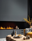 Modern indoor artificial fireplace with a black finish, featuring a visible water vapour flame effect in a stylishly decorated living room setting with a wooden coffee table, yellow sofa, and decorative items.