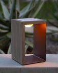 Modern outdoor solar lantern with wooden finish on a concrete surface, blurred green plants in the background by Les Jardins available at Spacio retail stores in India.