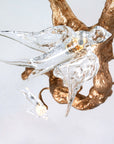 Close up of a chandelier with glass bird sculptures on a gold antler against a light gray background