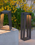 Two modern outdoor lamps on a stone ledge with yellow flowers in the background by Les Jardins available at Spacio retail stores in Mumbai & Kolkata.