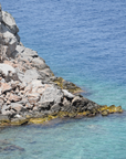 A person is standing on a rocky shore next to a body of water, experiencing the wellness brought by the calming Glamfusion Spilia from Glamora.