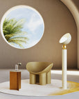 A versatile and vibrant lighting design from the Aurora Collection by Italamp featuring brushed gold accents, white glass diffuser, and retro-inspired finishes. An ivory finish floor lamp styled in a design set next to a chair with a round opening window showing tropical palm tree