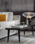 A table lamp on a marble side table next to a sofa with an orange pop colour cushion in a luxury living room with a dark theme. Cicla collection by Italamp, featuring handmade Balloton glass lamps in transparent, white, and smoked finishes with grooved Vicenza stone bases and metal accents.