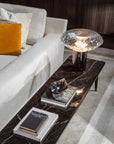 A table lamp on a marble long side table next to a sofa in a contemporary theme living room styled along with coffee table books and art objects. Cicla collection by Italamp, featuring handmade Balloton glass lamps in transparent, white, and smoked finishes with grooved Vicenza stone bases and metal accents.