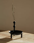 A black metal vase with a wide shallow bowl, three short legs, and a vertical stem with three round beads, holding dried stems, set on a light wood surface against a warm beige background.