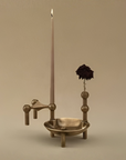 A vintage-style brass candle holder with a tall candle in a holder, a small bowl, and a dark dried flower, set against a solid beige background.