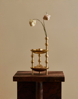 A small gold-toned decorative vase or candle holder with a geometric beaded frame, displayed on a dark wooden pedestal against a warm beige background. Two dried pale flowers are placed in the opening.