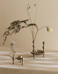 A styled tabletop scene with a small metallic vessel holding a dried flower stem, a clear glass bottle, and a small metallic object in the foreground, all set against a warm beige wall and draped cream fabric.