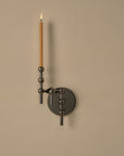 A modern wall-mounted candle holder with a circular dark metal backplate and an adjustable arm holding a single lit slim taper candle, shown against a plain beige wall.