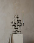 A black metal multi-arm candelabra sculpture with two lit slim LED taper candles, displayed on a white pedestal against a textured beige plaster wall.