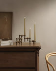 A three-tiered bronze candelabra holding three lit taper candles, styled on a dark wood chest of drawers in a warm, minimalist room.