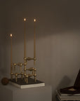 A brass three-arm candelabra with three lit taper candles, displayed on a white pedestal in a dimly lit room next to a small wooden box.
