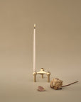A minimalist brass candle holder with a single lit white taper candle, styled with dried botanical elements on a warm beige background.