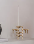 A modern brass candlestick holder with a tall white taper candle, styled on a white shelf next to a dark ceramic pot and a book against a neutral light gray wall.