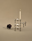 A brushed chrome metal candle holder with a lit white taper candle, displayed on a warm beige background next to a small clear glass dish holding dried dark flowers.