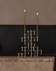 A sculptural metal candelabra with two lit taper candles, displayed on a stone mantel against a dark wood and beige wall background.